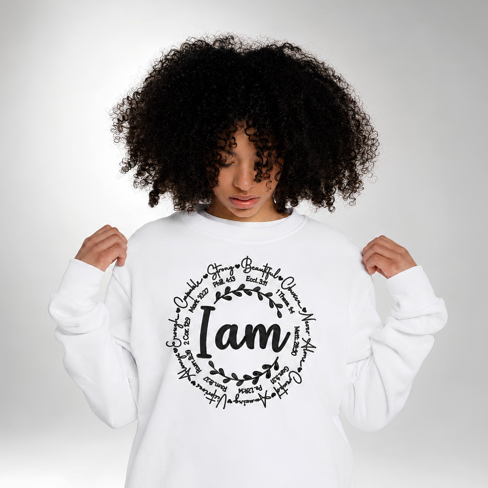 A woman with curly black hair wearing a white crewneck sweatshirt with a black graphic print that reads 'I am' surrounded by hand-written words and leafy branches, set against a light gray studio background.