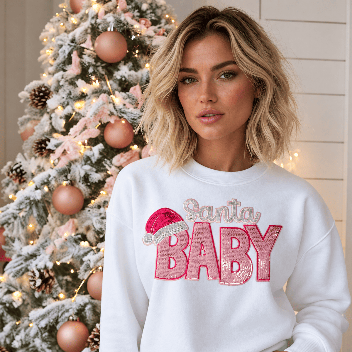 Digital mockup: woman by a frosted Christmas tree in a white crewneck with a pink sequin Santa Baby patch